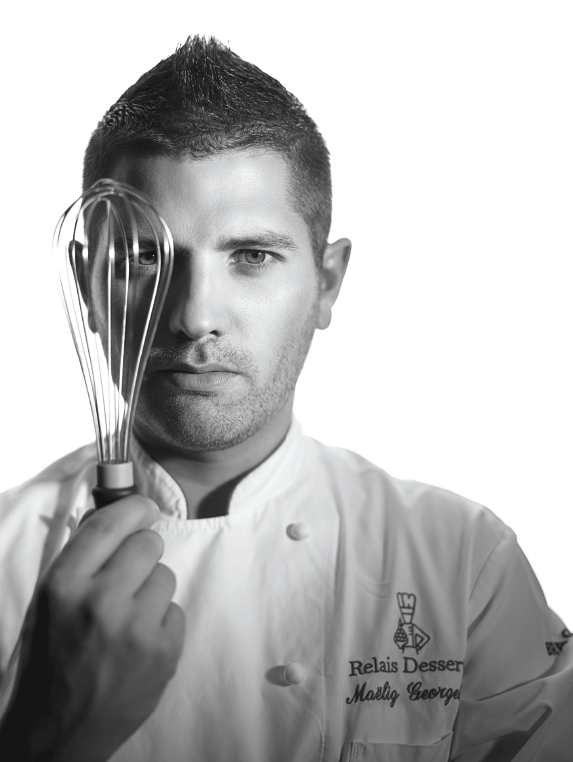 Le Chef Maëlig Georgelin pose avec un fouet à pâtisserie. Il porte un tablier Relais Desserts International.